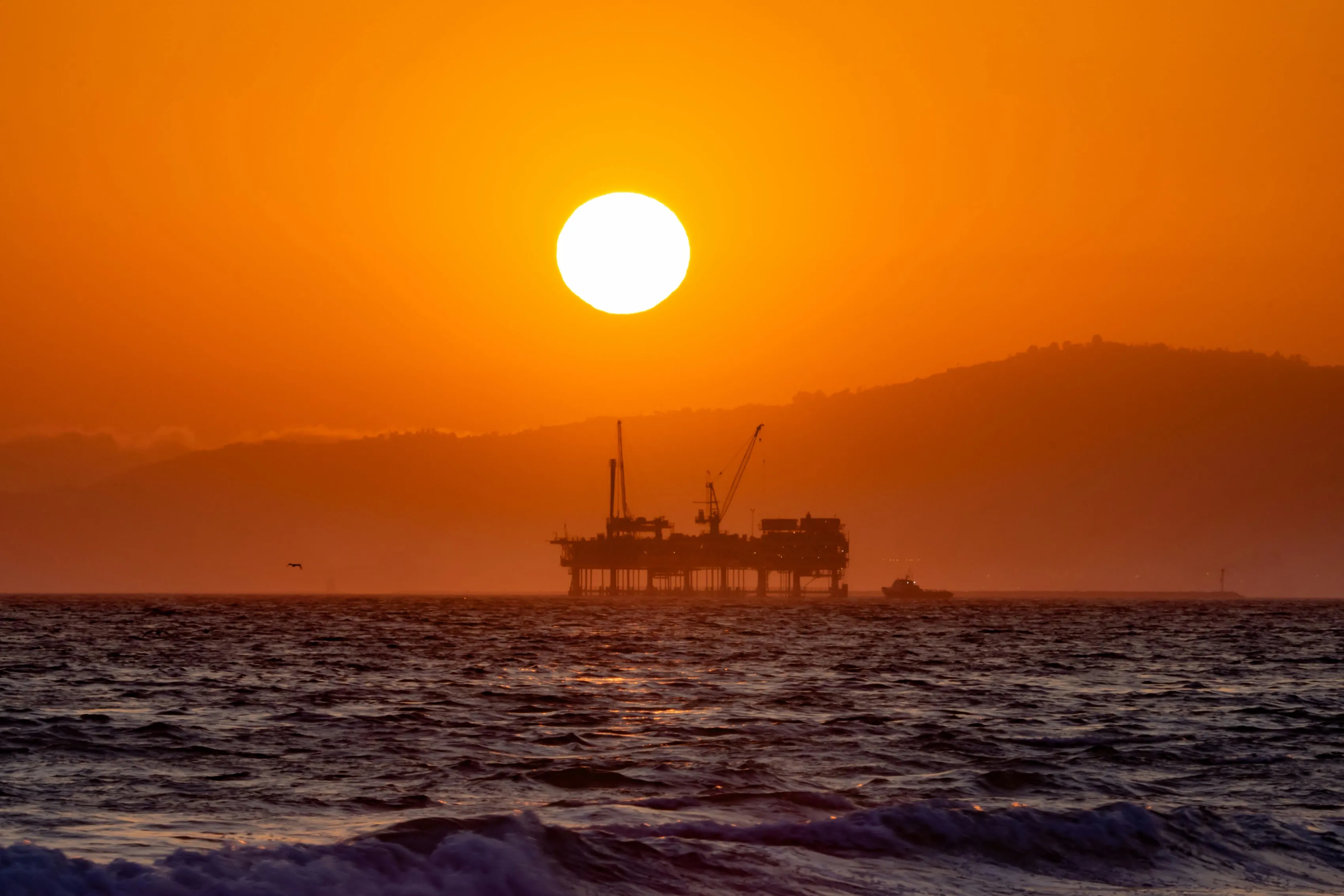 Offshore oil rig silhouetted against a vibrant sunset over the ocean, with waves in the foreground and a mountainous landscape in the background.